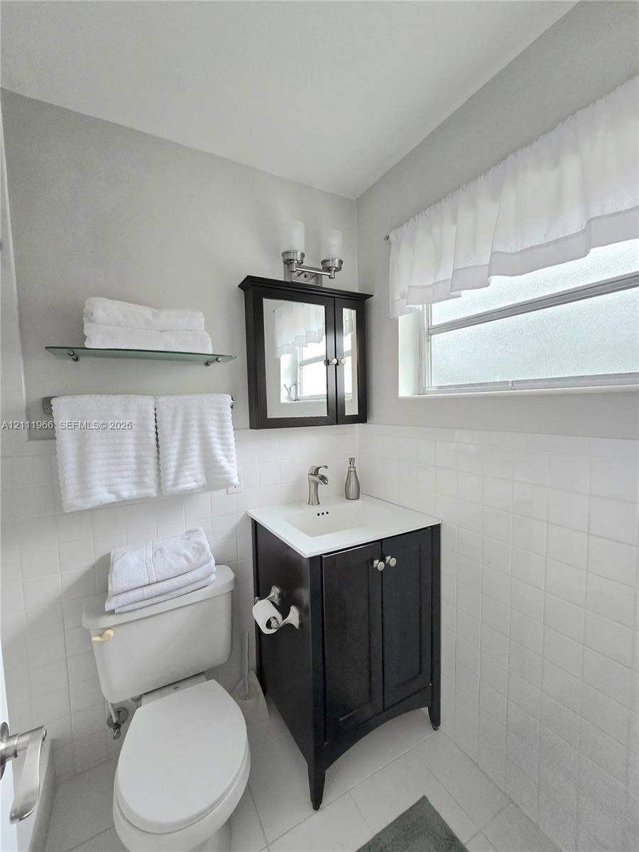 5387 SW 40th Avenue, #203, Dania Beach, FL 33314 Dark-finish wooden vanity with a white countertop and integrated sink, complemented by a matching mirrored medicine cabinet, a wall-mounted glass shelf, and classic white tiled wall surfaces