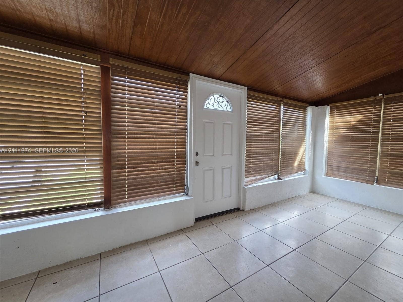 600 SW 44th Court, Miami, Miami, FL 33134 Enclosed porch featuring wood-paneled ceiling, large-format tile flooring, multiple windows with fitted blinds, and a white entry door with a decorative arched transom window
