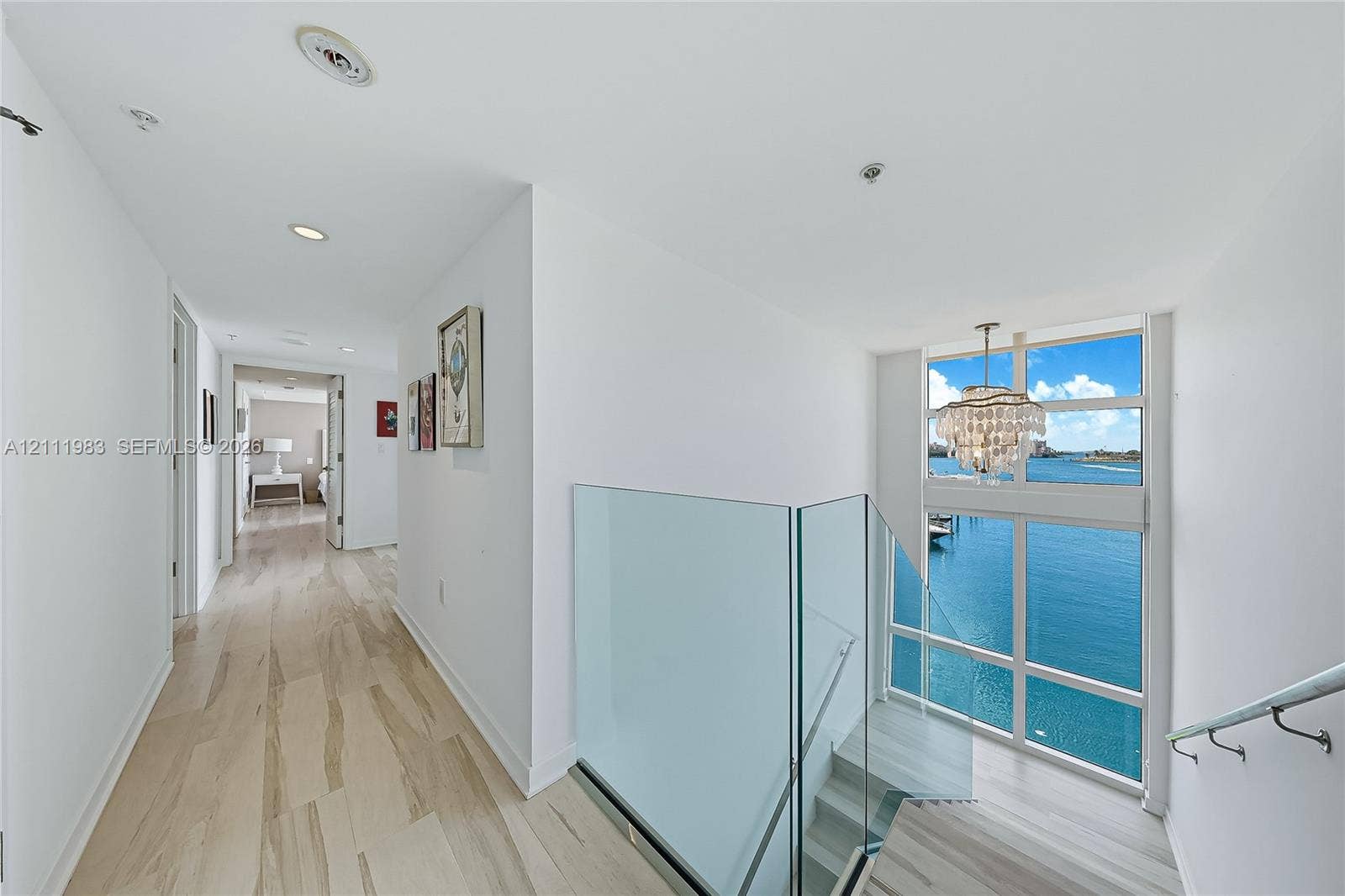 90 Alton Road, #TH-8, Miami Beach, FL 33139 Upstairs hallway and staircase featuring light-toned wood-finish flooring, glass stair railings, multi-panel floor-to-ceiling windows, and a decorative chandelier