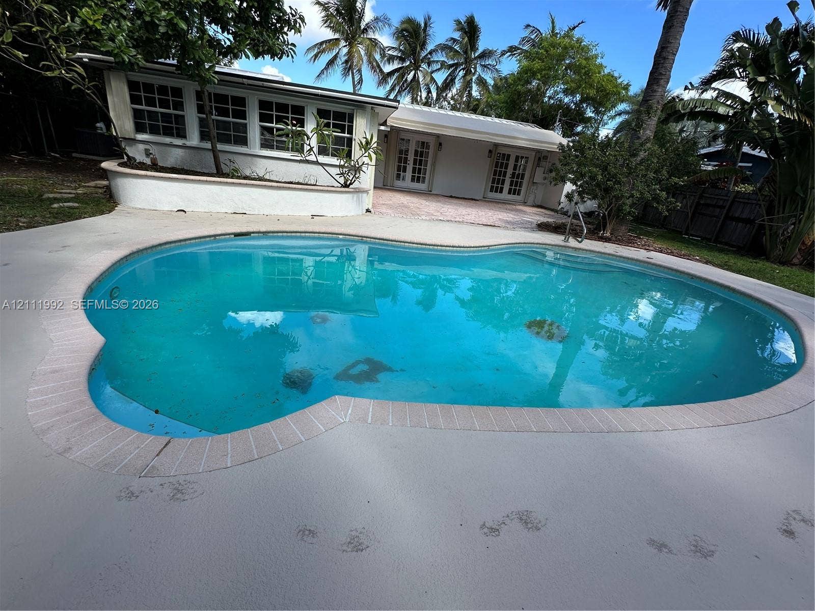 In-ground swimming pool with brick-style coping, concrete patio, white exterior walls, and dual sets of French doors