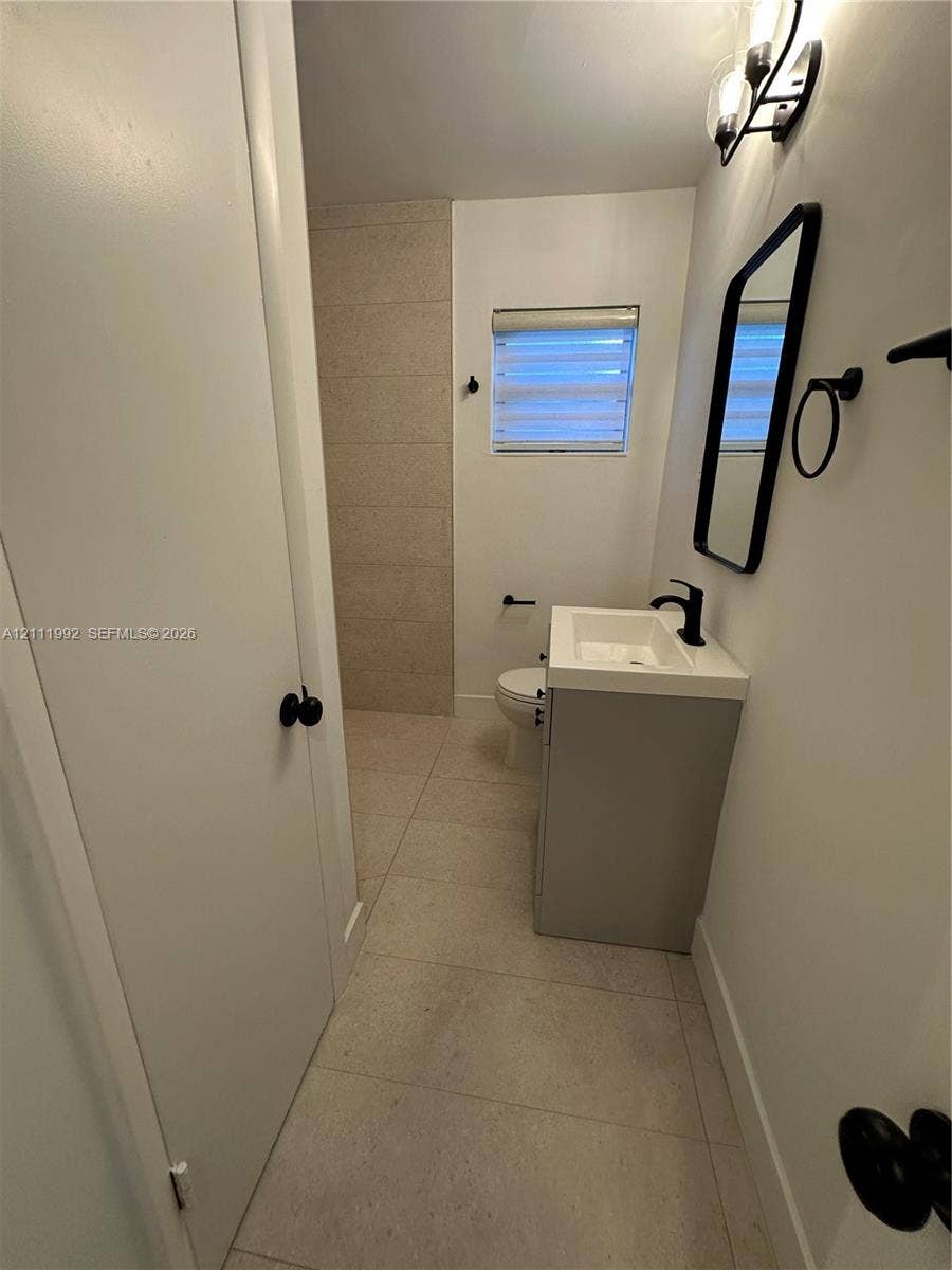Bathroom featuring large-format neutral tile flooring, a modern grey vanity with a white rectangular sink, matte black hardware, and a framed vanity mirror