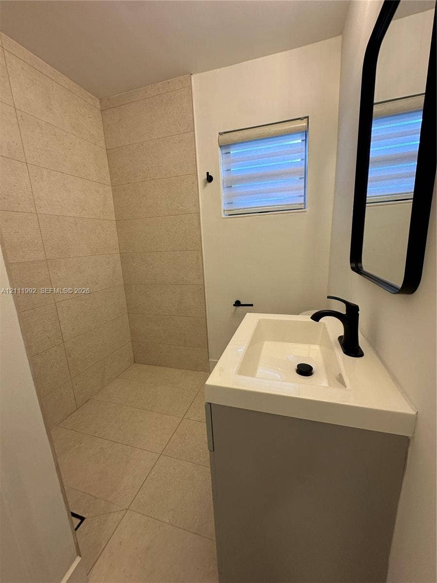 Walk-in shower with floor-to-ceiling neutral tile, modern vanity with a rectangular sink, matte black faucet, wall-mounted mirror with a black frame, and window with integrated blinds
