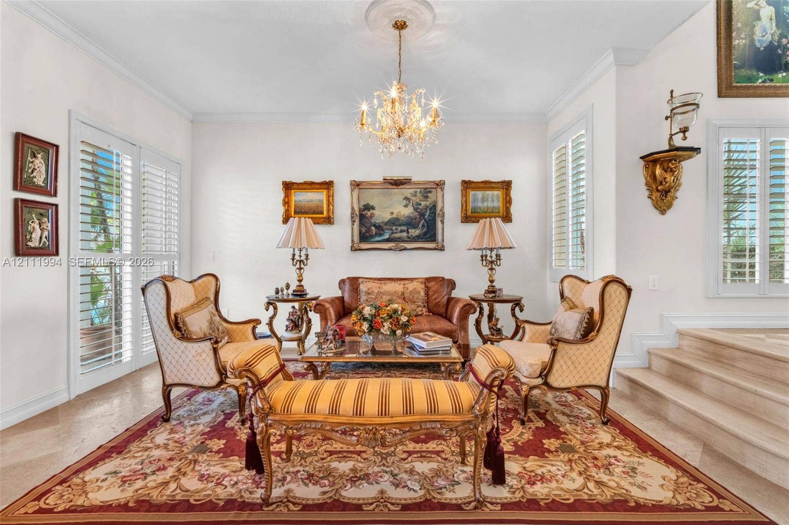 Formal living area featuring polished stone flooring, custom plantation shutters, tiered stone steps, crown molding, and a central crystal chandelier with a decorative ceiling medallion