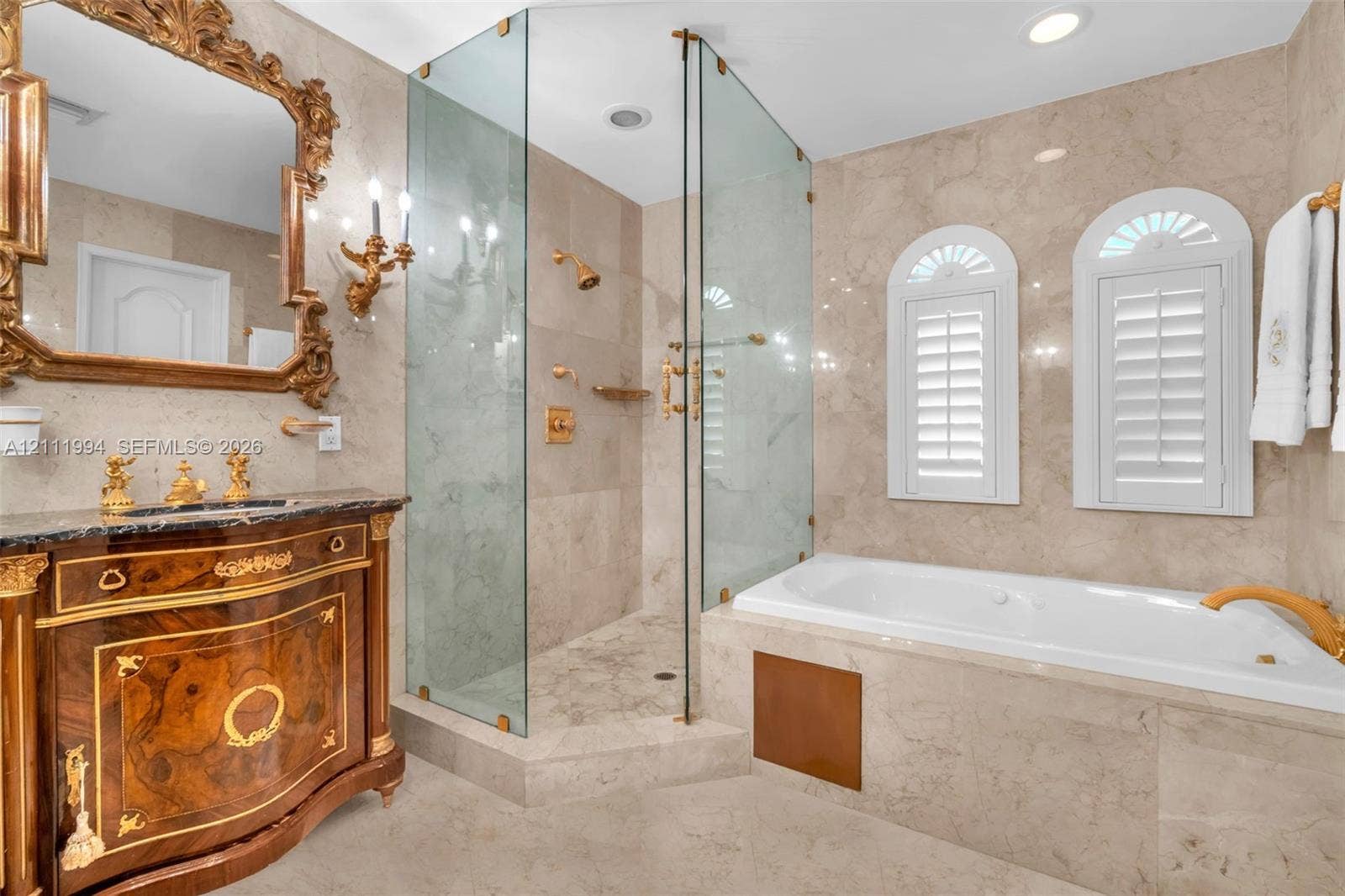 Bathroom featuring full-height natural stone tiling, a glass-enclosed shower, a built-in soaking tub, a custom vanity with gold-tone hardware, and arched window shutters