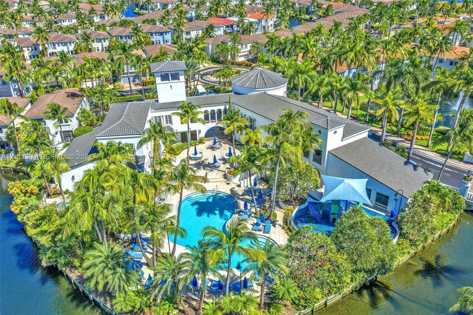 Community clubhouse featuring a tiled roof, architectural tower, swimming pool, water-feature area, and surrounding palm trees