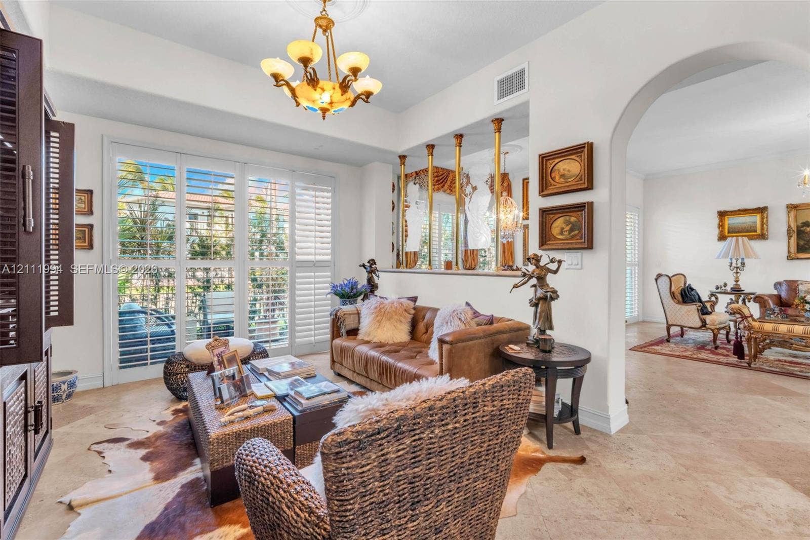 Living space featuring large-format tile flooring, arched entryways, plantation shutters, a decorative half-wall with brass-tone pillars, and a multi-arm chandelier