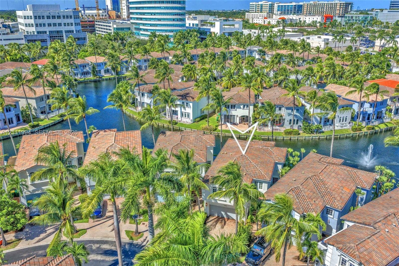 Aerial perspective of a multi-story residence featuring a terracotta-tiled roof, concrete exterior, attached two-car garage, and waterfront positioning along a canal with manicured landscaping and mature palm trees