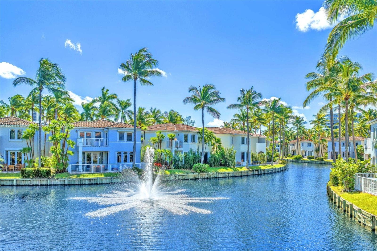 Waterfront residences featuring tile roofs, private balconies, a central water fountain, and mature palm trees