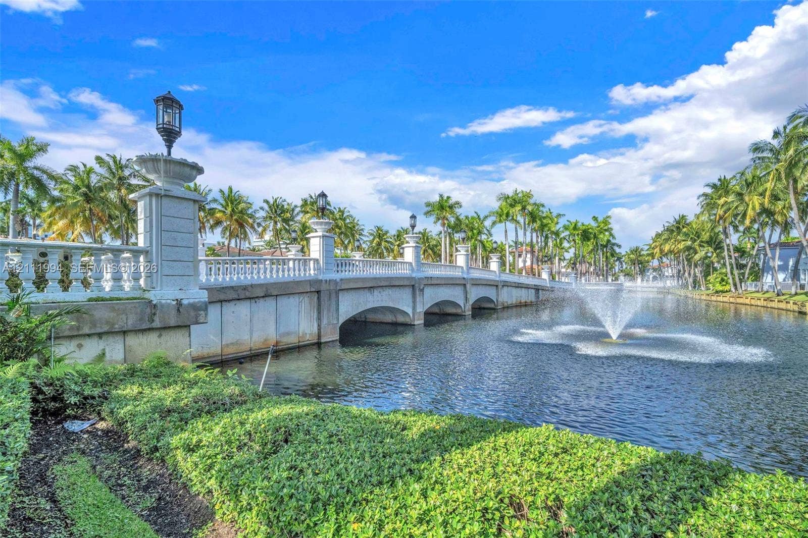 Stone arched bridge featuring white balustrades, decorative post-top lighting, water fountain, and mature palm tree landscaping