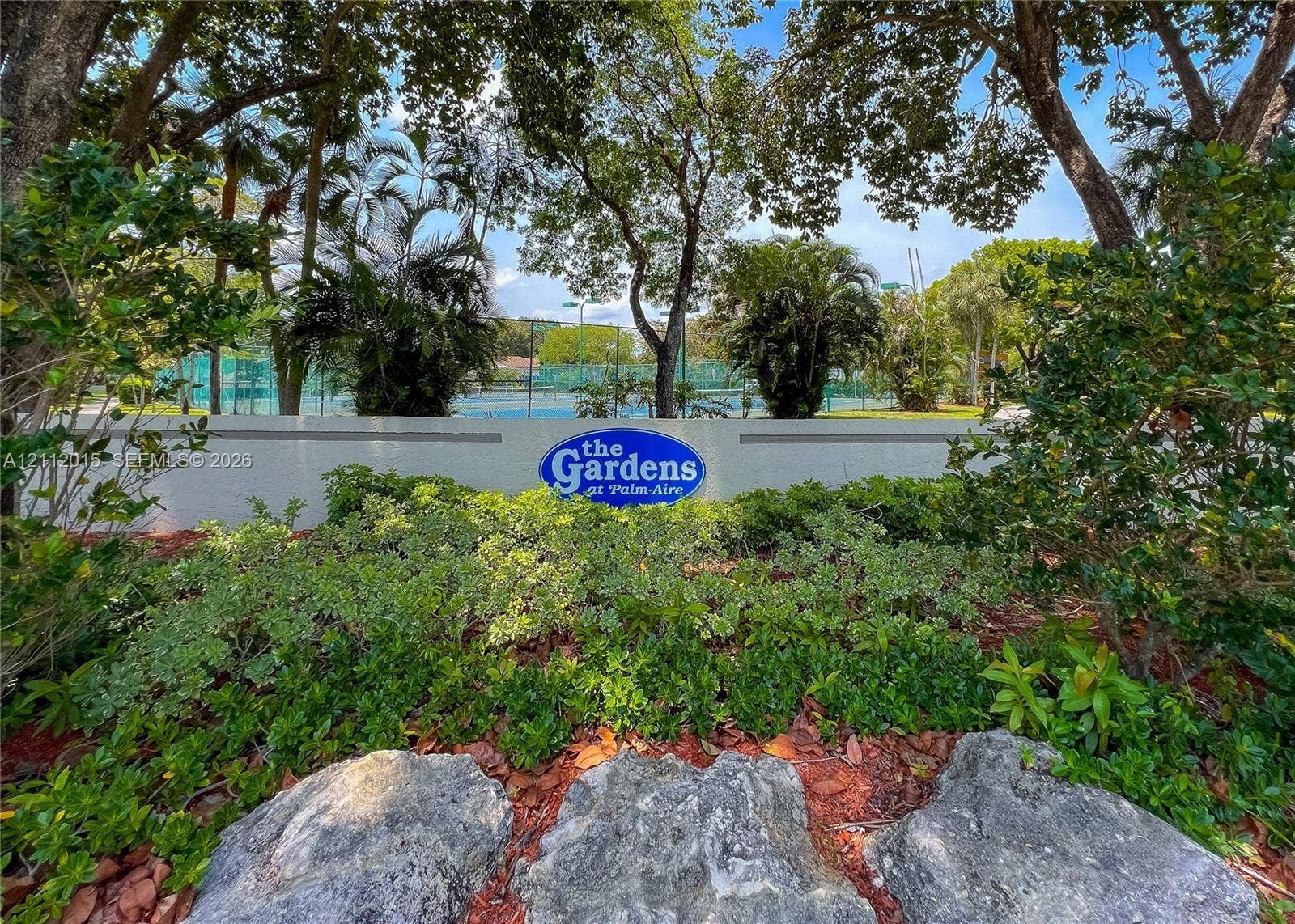 717 Gardens Drive, #201, Pompano Beach, Pompano Beach, FL 33069 Landscaped entrance featuring mature shade trees, stone boulders, shrubbery, and signage for The Gardens at Palm-Aire