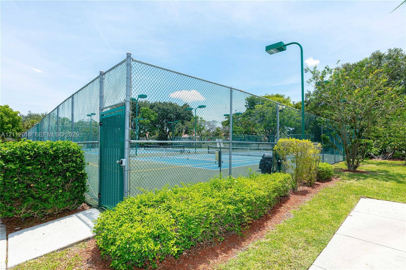 717 Gardens Drive, #201, Pompano Beach, Pompano Beach, FL 33069 Tennis court enclosed by chain-link fencing, featuring overhead lighting, a gated entrance, and manicured landscaping