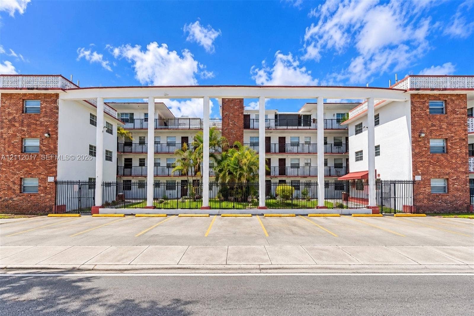 Multi-story exterior featuring brick masonry, white structural pillars, integrated balconies, and perimeter security fencing