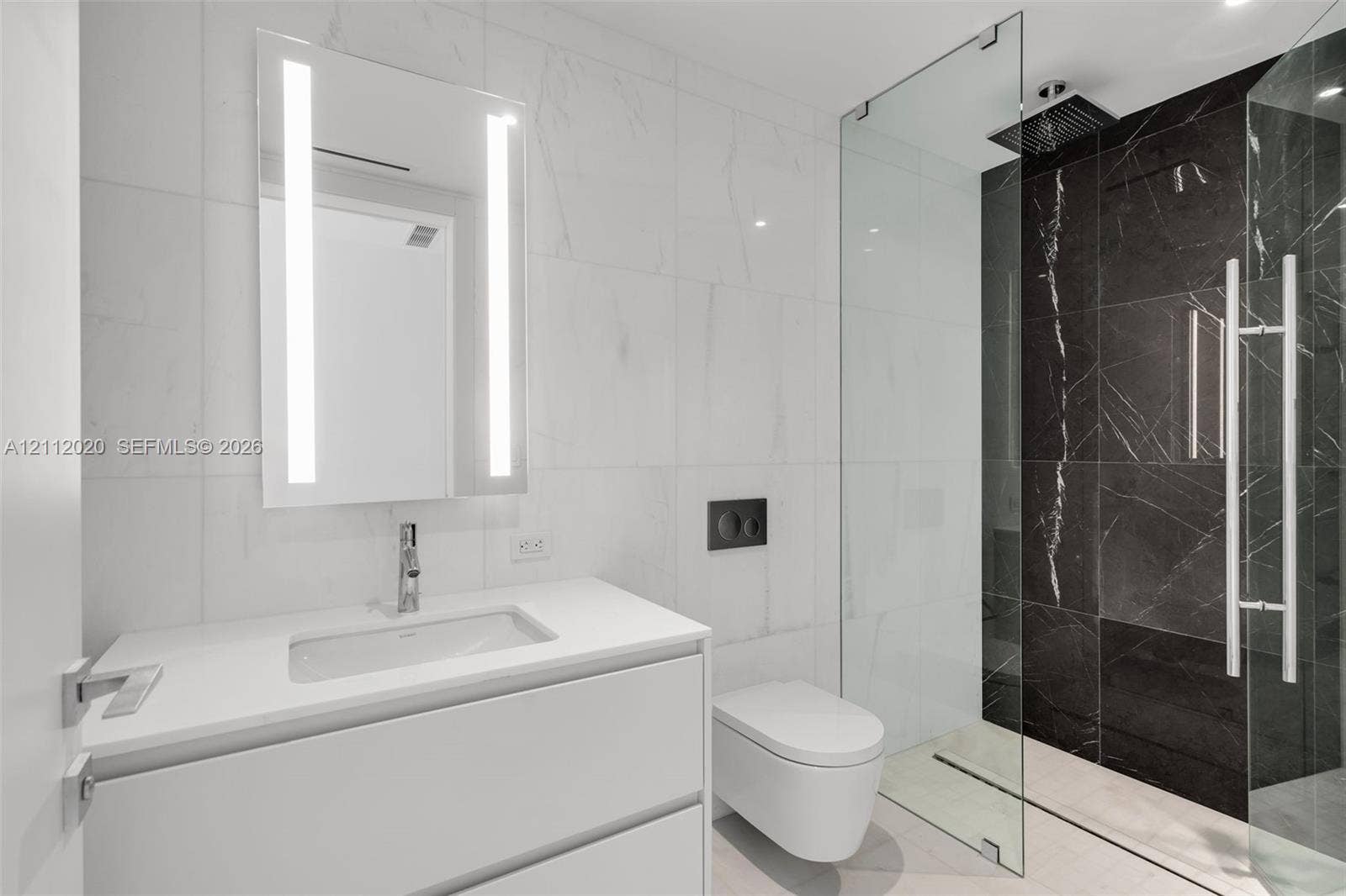 300 Biscayne Blvd Way, #4202, Miami, Miami, FL 33131 Wall-mounted vanity with a white countertop and rectangular undermount sink, illuminated vertical mirror, wall-hung toilet with a dual-flush actuator, and walk-in shower featuring a glass enclosure and dark tile surround