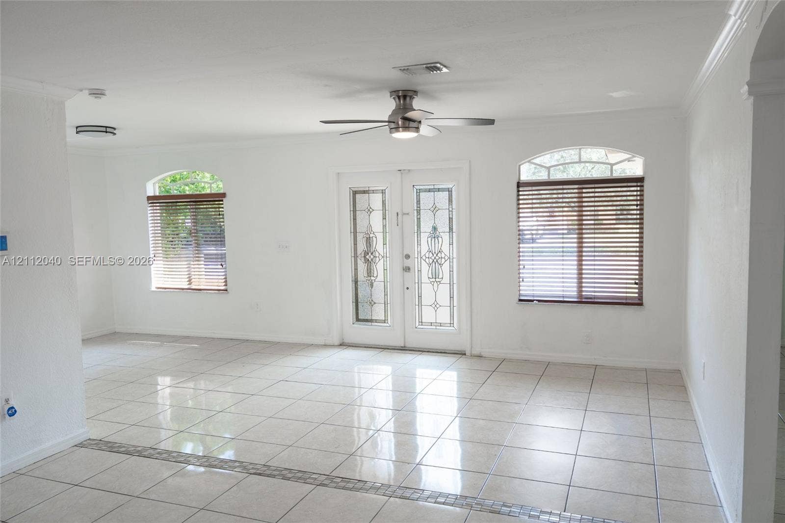 Open-concept interior featuring light-toned tile flooring, double entry doors with decorative glass inserts, arched windows, and a ceiling fan with integrated lighting