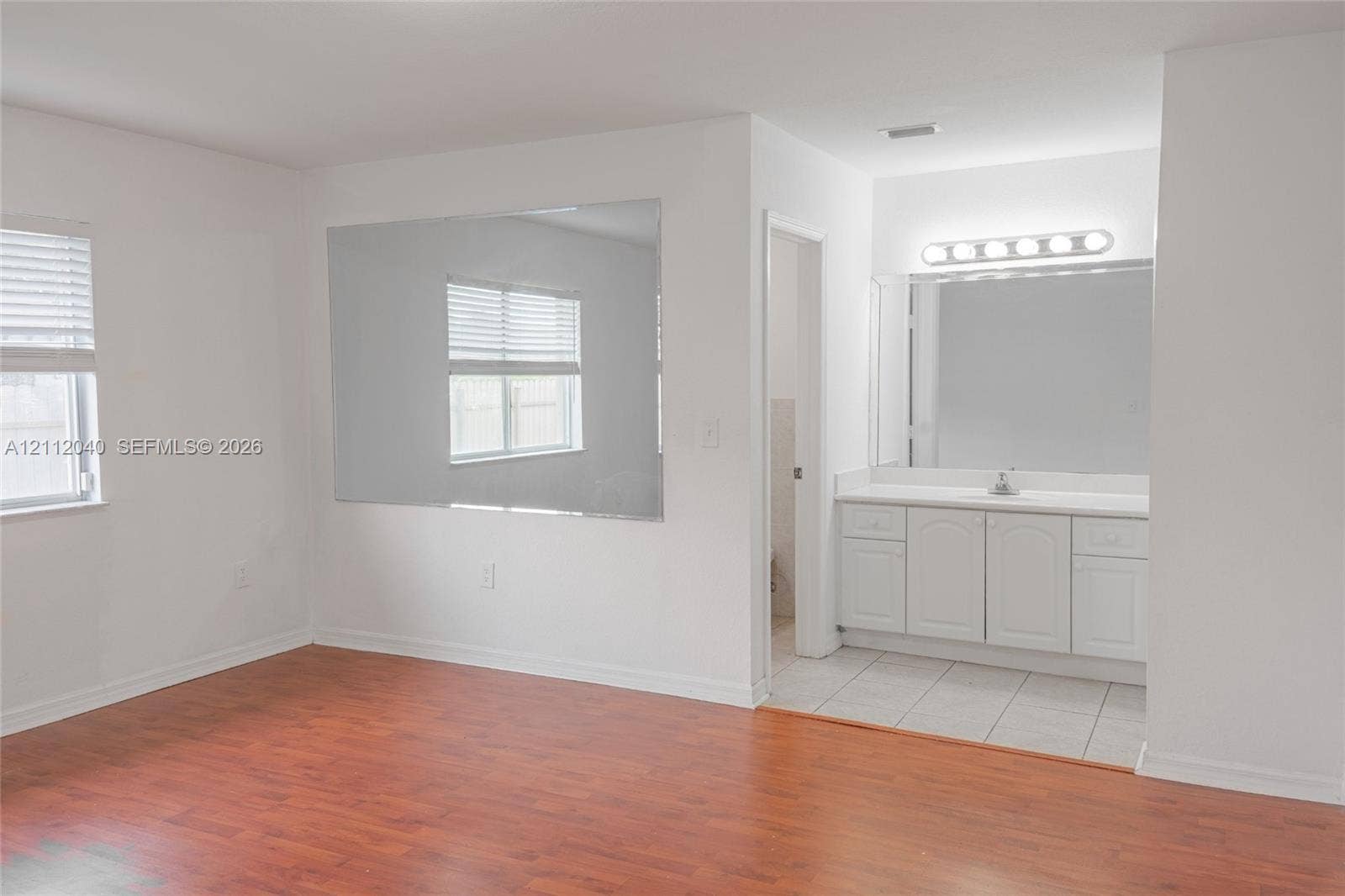 Wood-finish flooring, white cabinetry with integrated countertop, large wall-mounted mirror, and horizontal window blinds