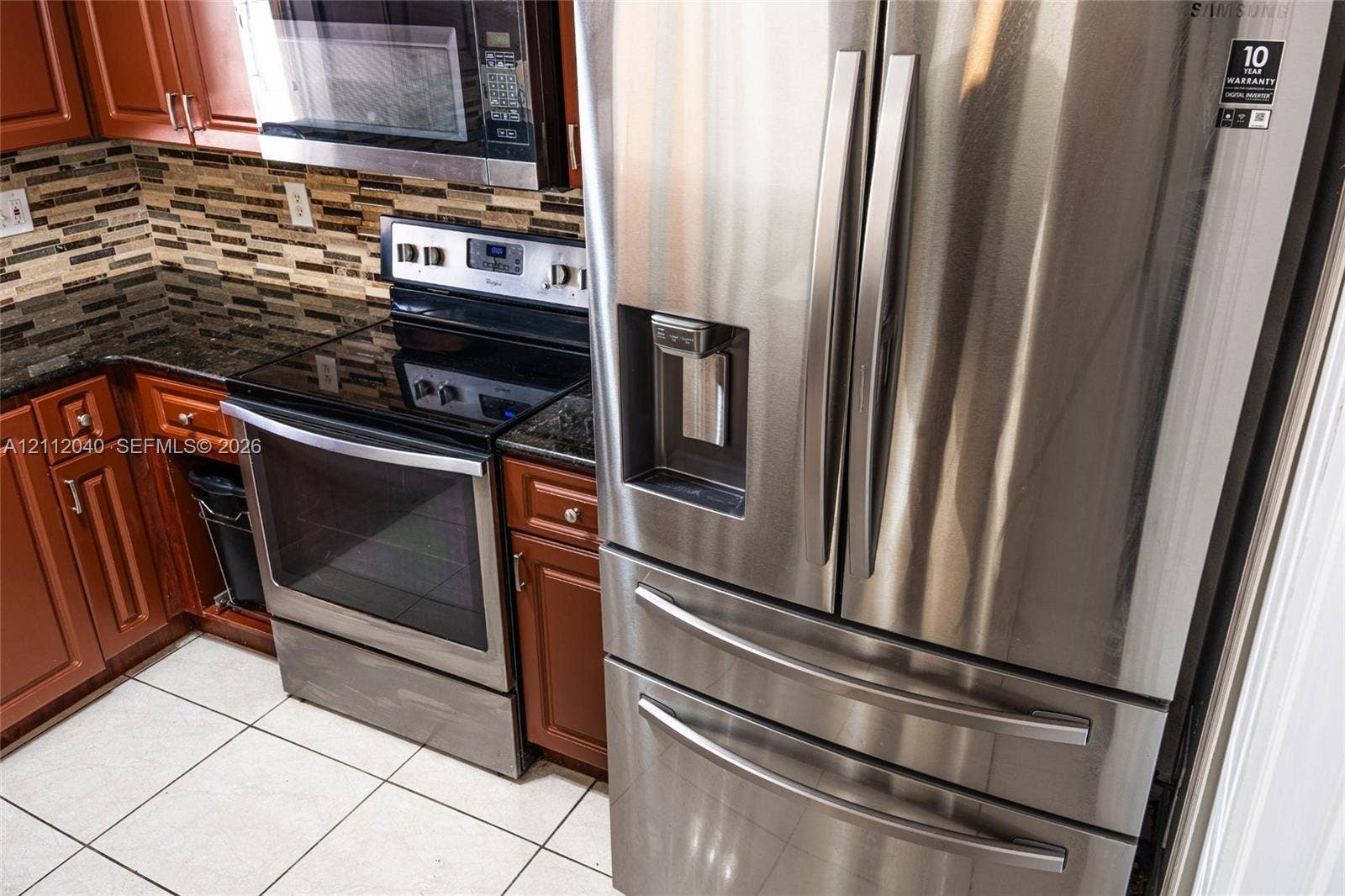 Kitchen featuring stainless steel appliances, dark stone countertops, natural wood-finish cabinetry, mosaic tile backsplash, and ceramic tile flooring