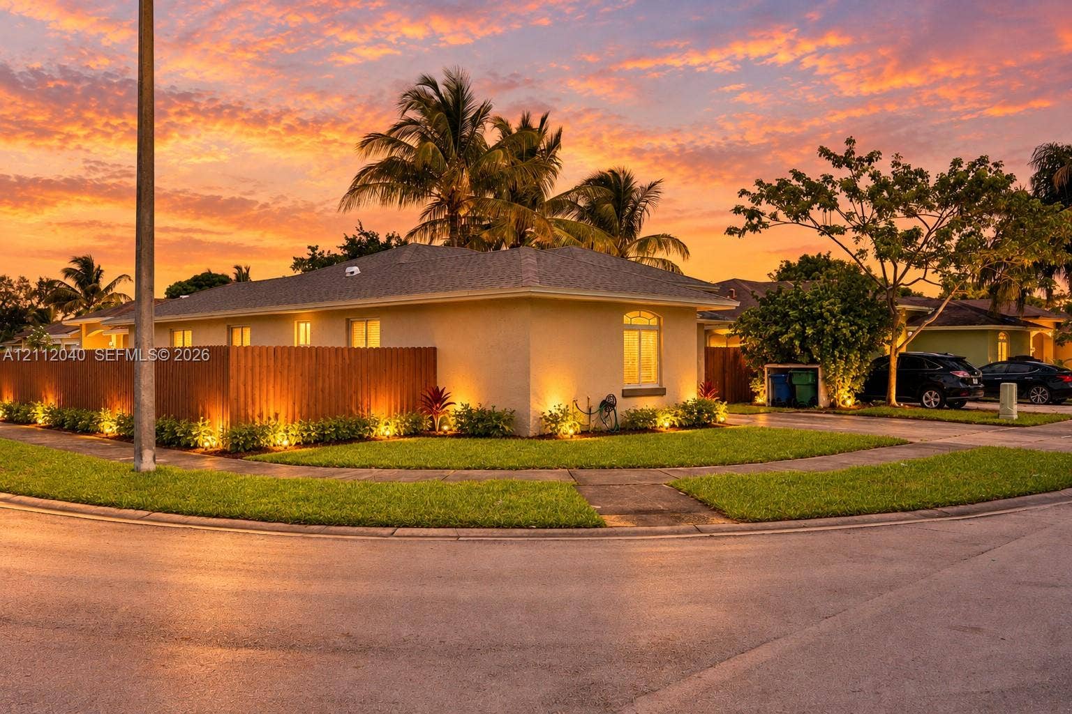 Stucco exterior residence featuring a tall wood privacy fence, integrated landscape lighting, manicured lawn, and mature palm trees