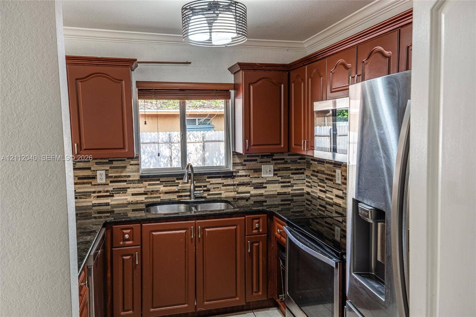 Kitchen featuring stained wood cabinetry, stone countertops, multi-toned tile backsplash, stainless steel appliances, and a circular drum pendant light fixture