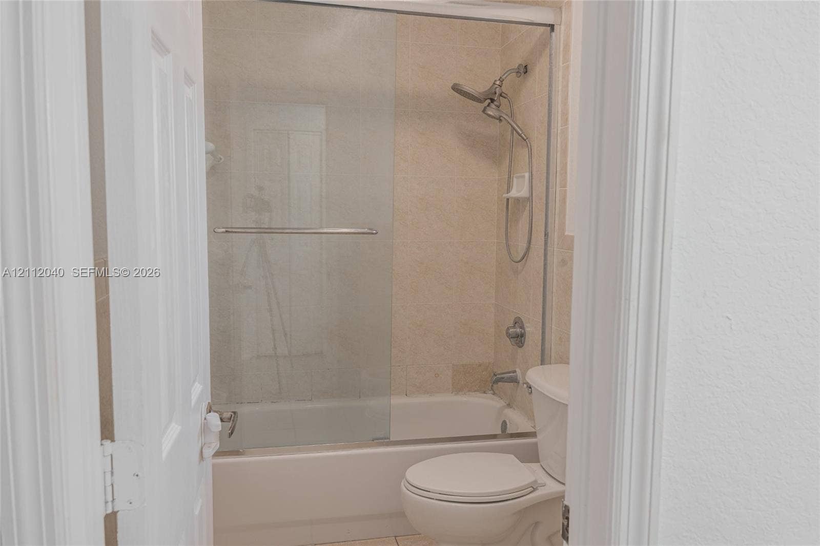 Bathroom featuring a tub-shower combination with sliding glass doors, neutral tile surround, adjustable handheld shower fixture, and a white toilet