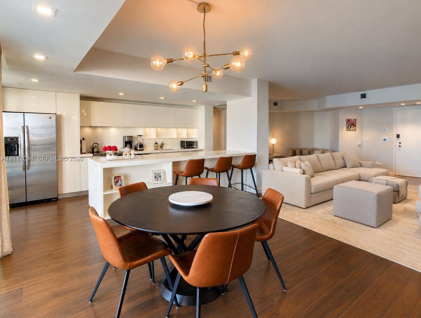 Open-concept kitchen featuring high-gloss cabinetry, stainless steel refrigerator, breakfast bar with waterfall-edge countertop, recessed lighting, and modern multi-arm chandelier