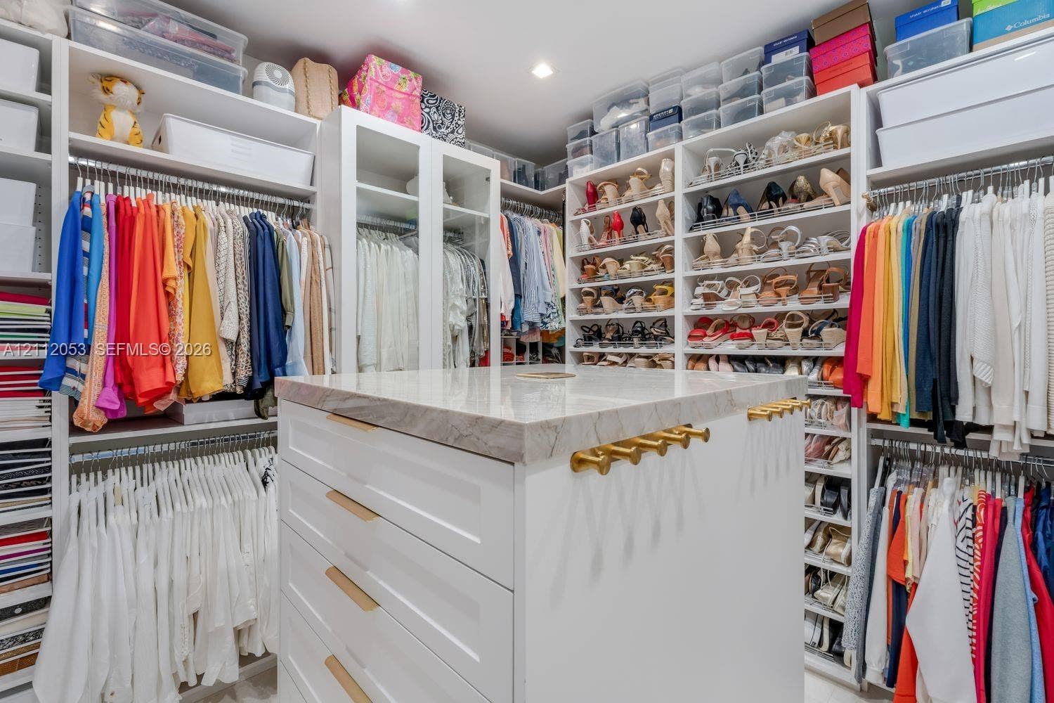 Custom walk-in closet featuring extensive built-in white cabinetry, open shelving, integrated hanging rods, and a center island topped with a stone-finish counter and gold-tone hardware