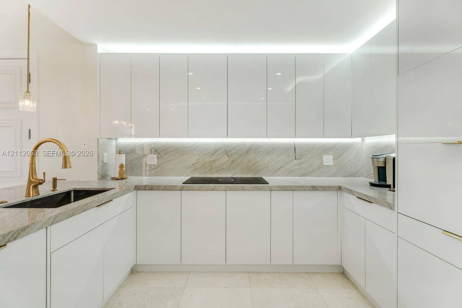 High-gloss white flat-panel cabinetry, stone countertops and backsplash with veined detailing, gold-tone faucet, under-cabinet lighting, and integrated cooktop