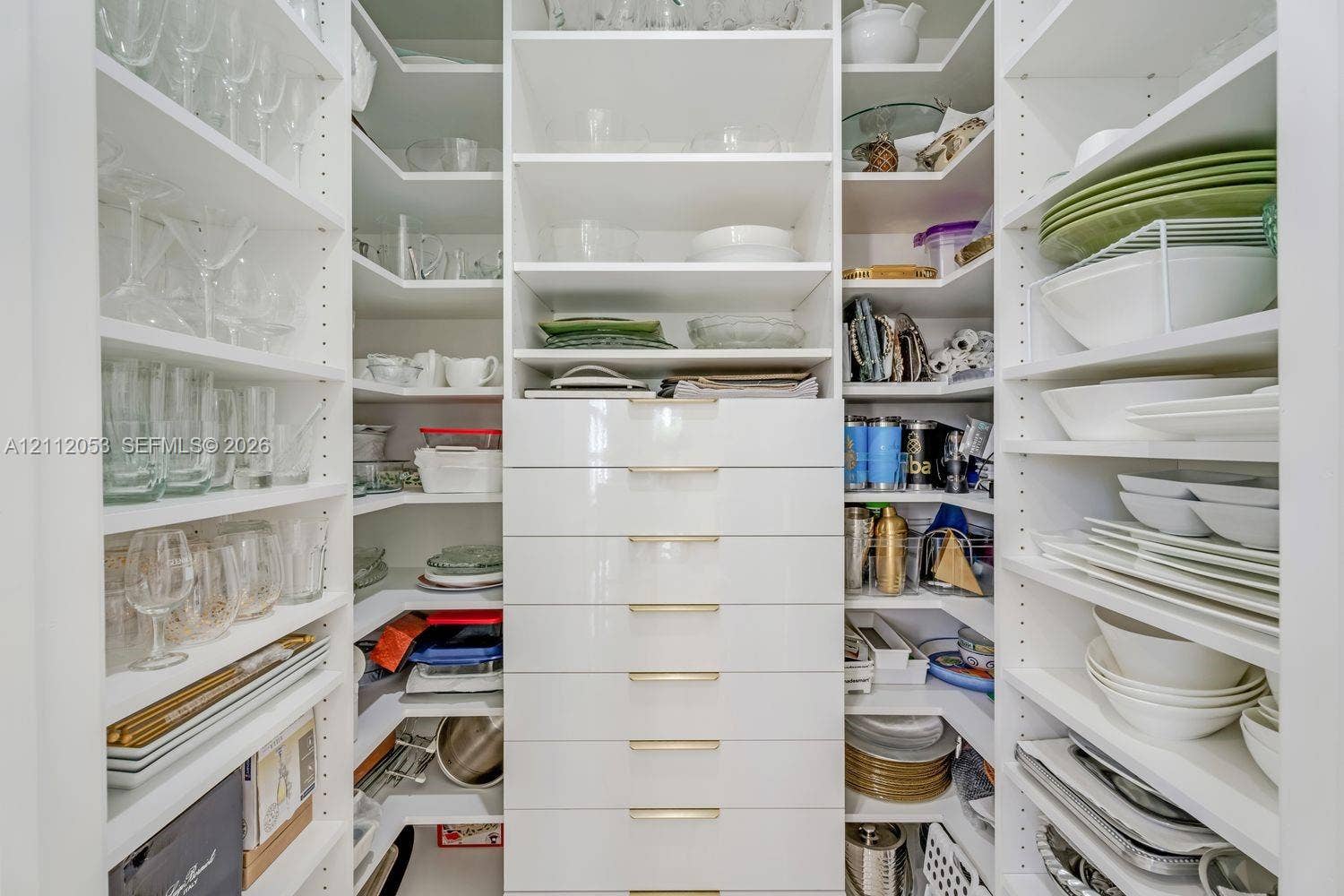 Custom walk-in pantry featuring white floor-to-ceiling shelving, tiered corner storage, and a central stack of drawers with metallic hardware