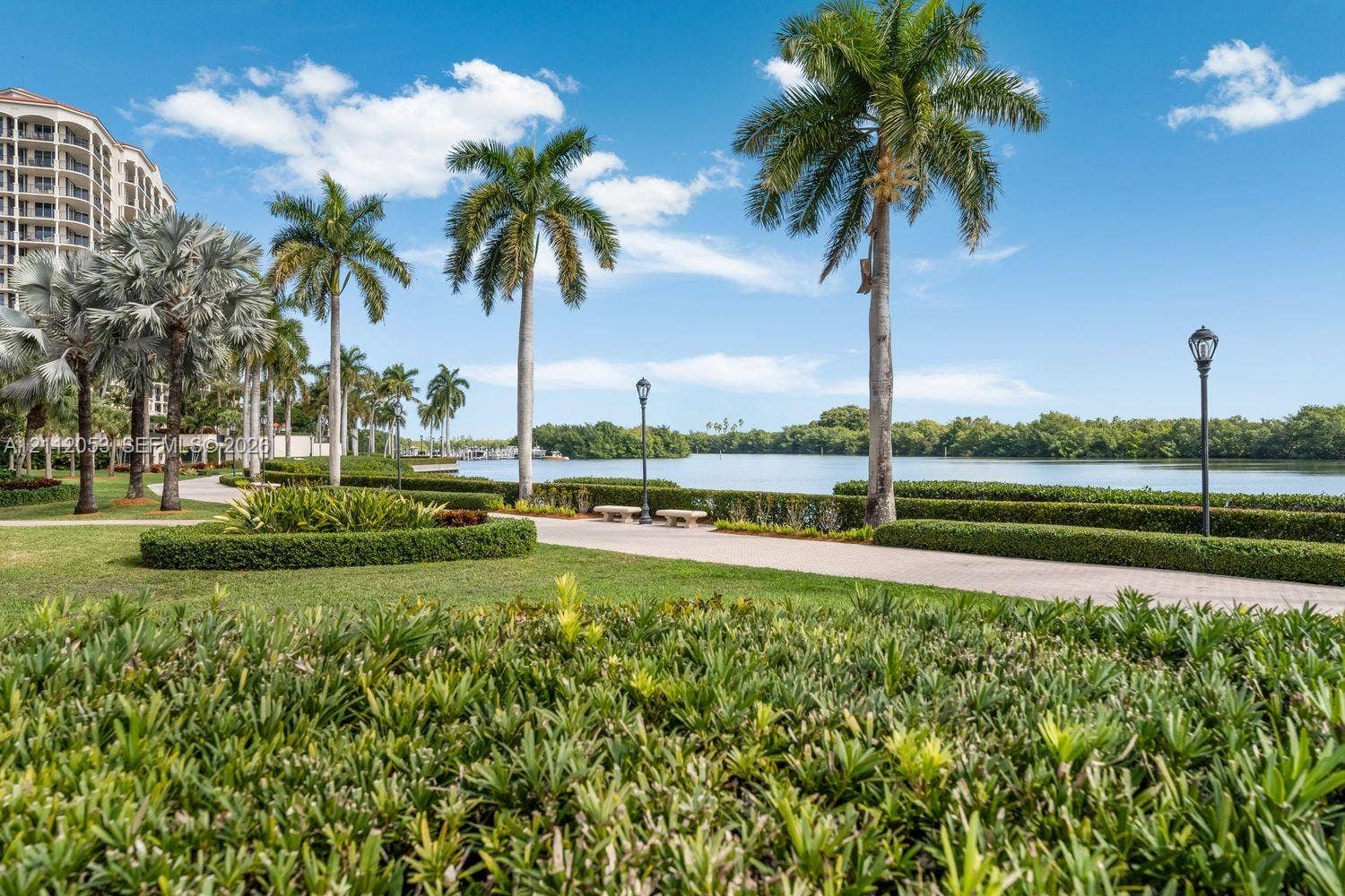 Waterfront grounds featuring manicured hedges, mature palm trees, paved walking paths, and ornamental lamp posts