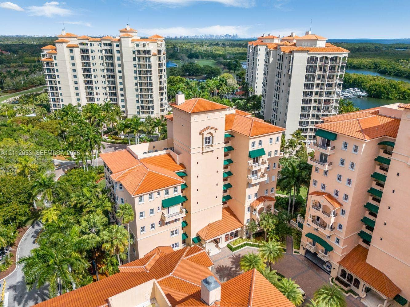 Aerial perspective of mid-rise residential structures featuring terracotta-style roof tiles, light-toned stucco facades, integrated balconies with green awnings, and landscaped grounds with mature palm trees