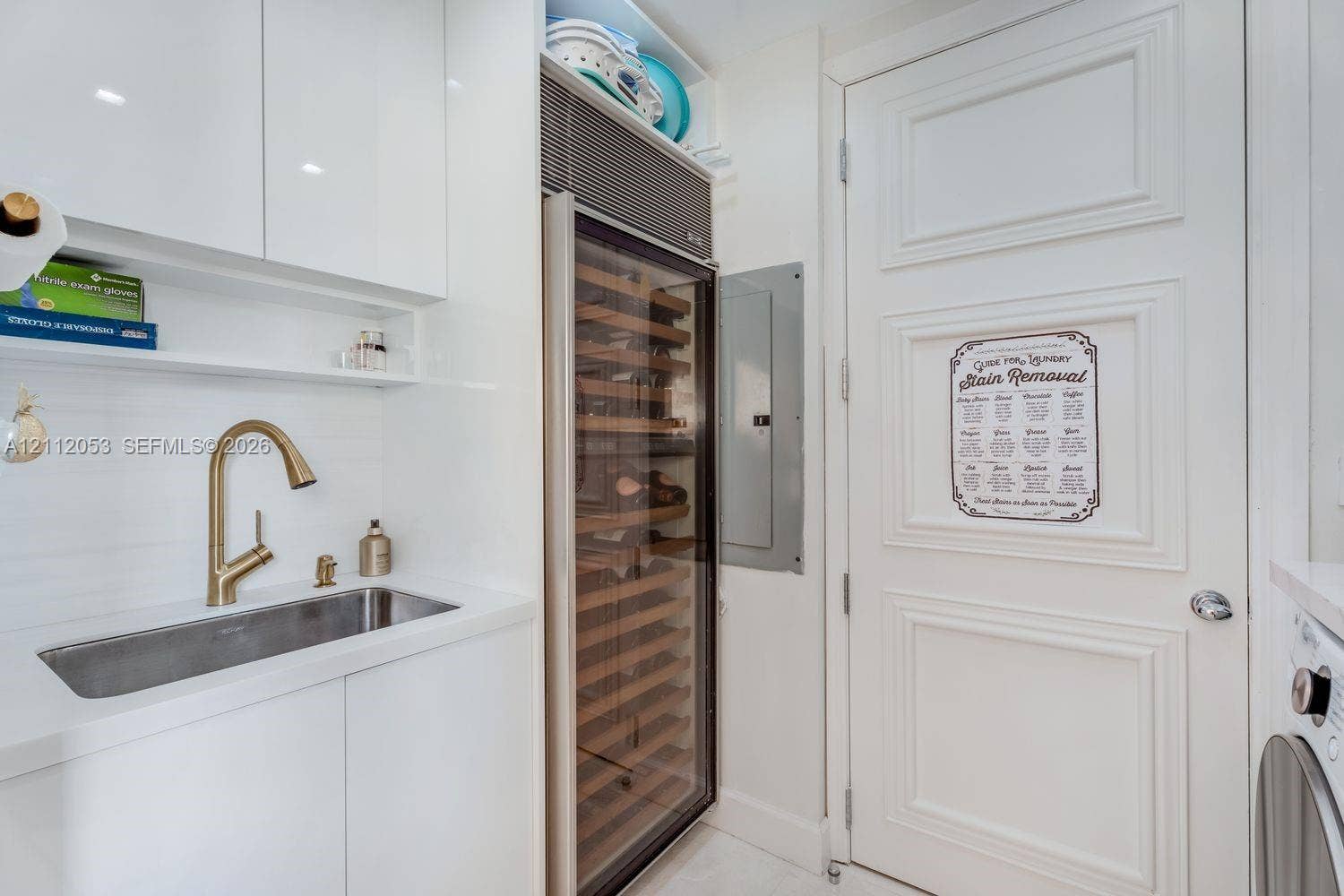 Built-in wine refrigerator, brass-tone gooseneck faucet, integrated stainless steel sink, custom white cabinetry, and multi-panel interior door
