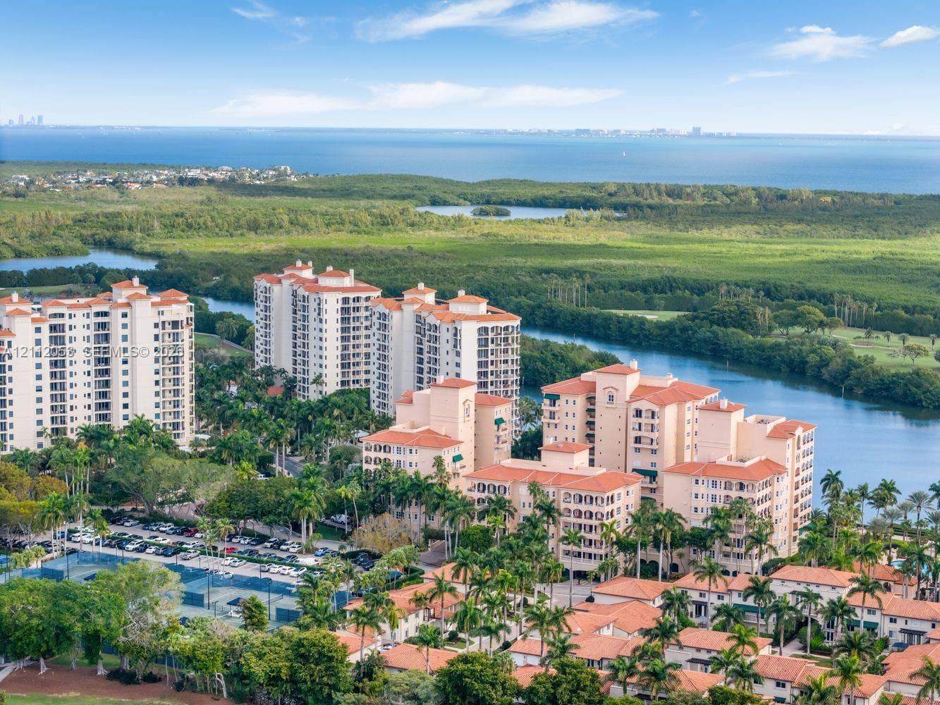 Aerial perspective of high-rise condominium complex featuring terracotta-tiled roofs, manicured palm landscaping, on-site tennis courts, and expansive water frontage