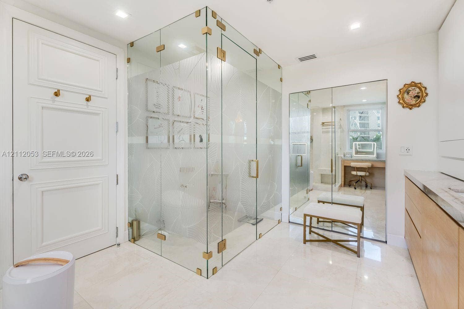 Frameless glass shower enclosure with gold-tone hardware, polished stone-finish flooring, custom light-wood cabinetry, and full-length mirrored panels