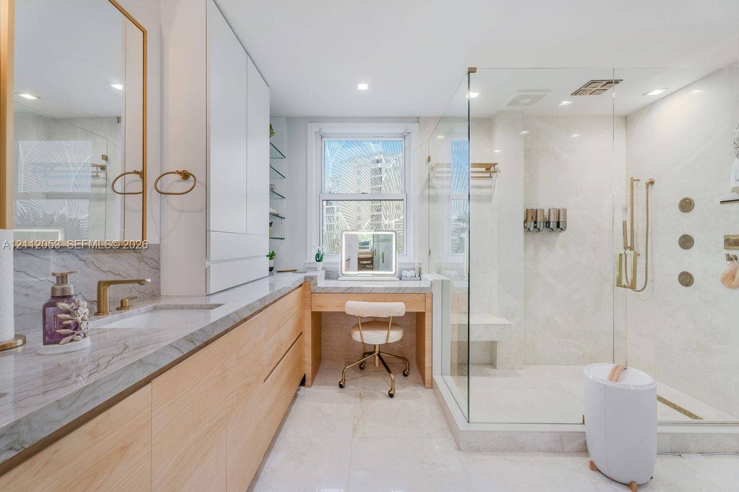 Bathroom featuring a light-wood vanity with stone-finish countertops, glass-enclosed walk-in shower with body sprayers and built-in bench, integrated custom cabinetry, and brass-finish fixtures