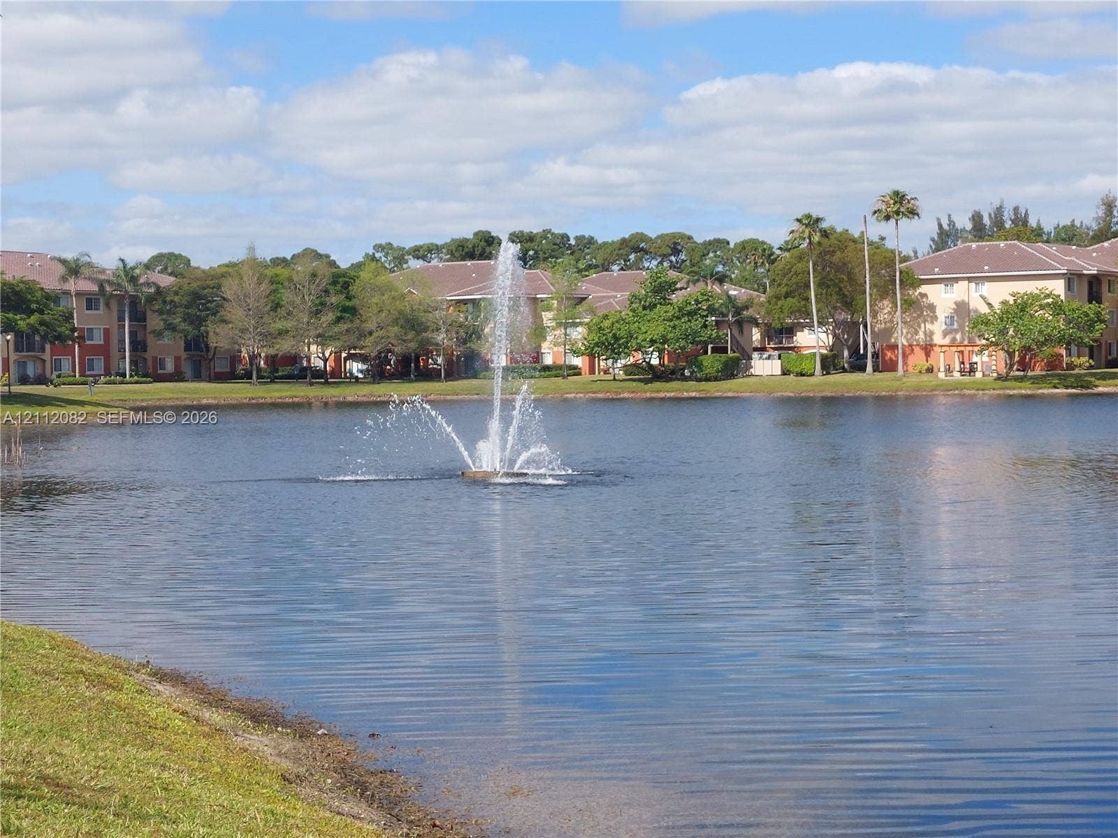 Waterfront setting featuring a central pond fountain, manicured lawn, and mature palm trees