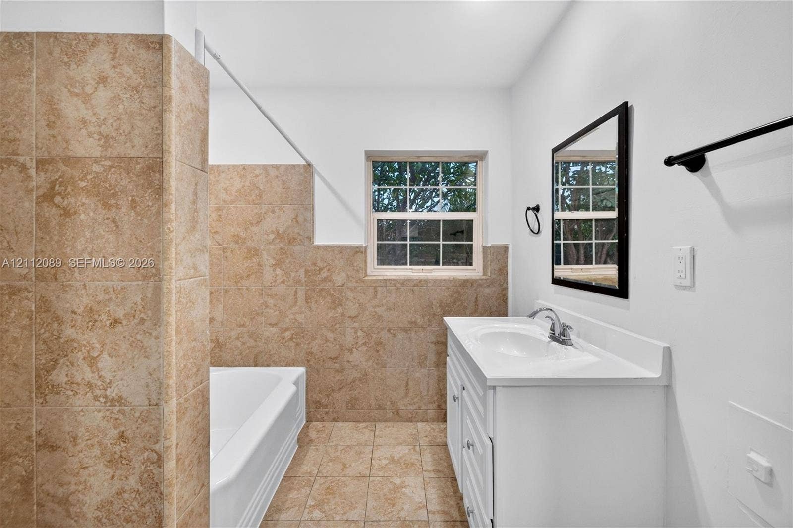 2017 NE 29th Court, Fort Lauderdale, FL 33306 Bathroom featuring a white vanity with an integrated sink, a deep soaking tub with a tiled surround, neutral floor tiling, a framed wall mirror, and matte black fixtures