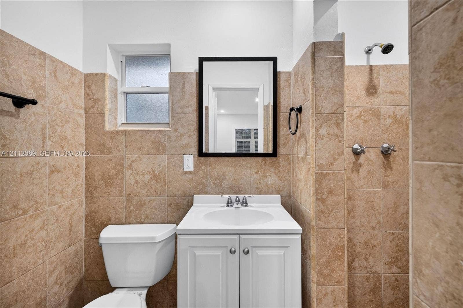 2017 NE 29th Court, Fort Lauderdale, FL 33306 Bathroom featuring tan stone-tile wall cladding, a white pedestal vanity with an integrated sink, a framed wall mirror, and a wall-mounted shower head