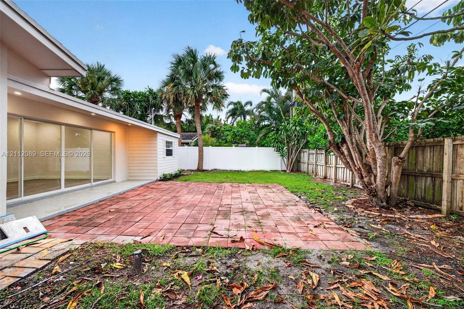 2017 NE 29th Court, Fort Lauderdale, FL 33306 Backyard featuring a red brick patio, white vinyl privacy fencing, mature trees, and glass sliding door access