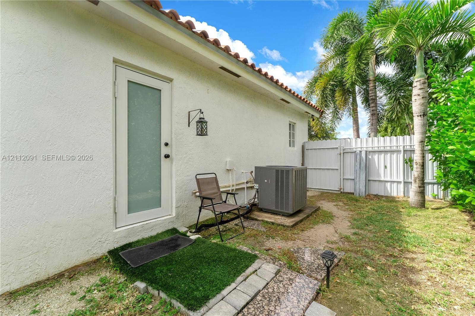 29481 SW 193rd Court, Homestead, FL 33030 Exterior side yard featuring white stucco siding, a glass-panel door, terracotta roof tiles, a white vinyl privacy fence, and mature palm trees