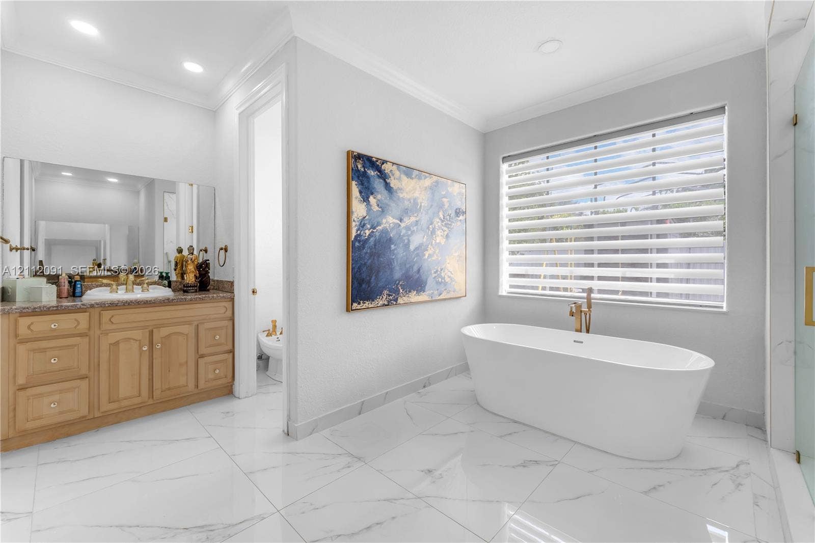 29481 SW 193rd Court, Homestead, FL 33030 Freestanding soaking tub with gold-tone fixtures, wood cabinetry with stone-surface countertop, white tile flooring with veining detail, and large-format wall mirror