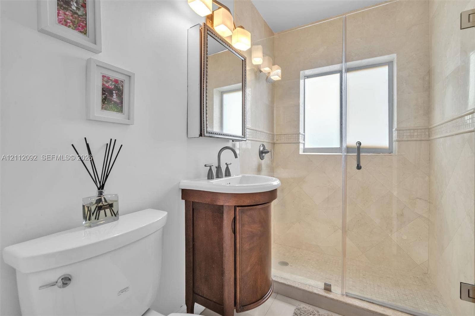 419 Sevilla Avenue, Coral Gables, FL 33134 Curved wood-finish vanity with a white basin, stone-tiled shower with a glass enclosure, and wall-mounted three-light fixture