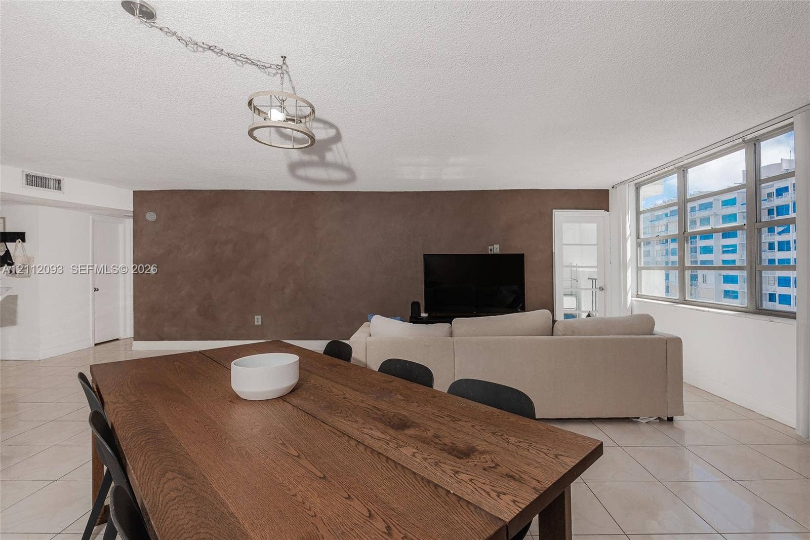 2750 NE 183rd Street, #1612, Aventura, FL 33160 Open-concept living space featuring large-format ceramic tile flooring, a prominent accent wall with textured finish, expansive multi-pane windows, and a suspended industrial-style drum pendant light fixture