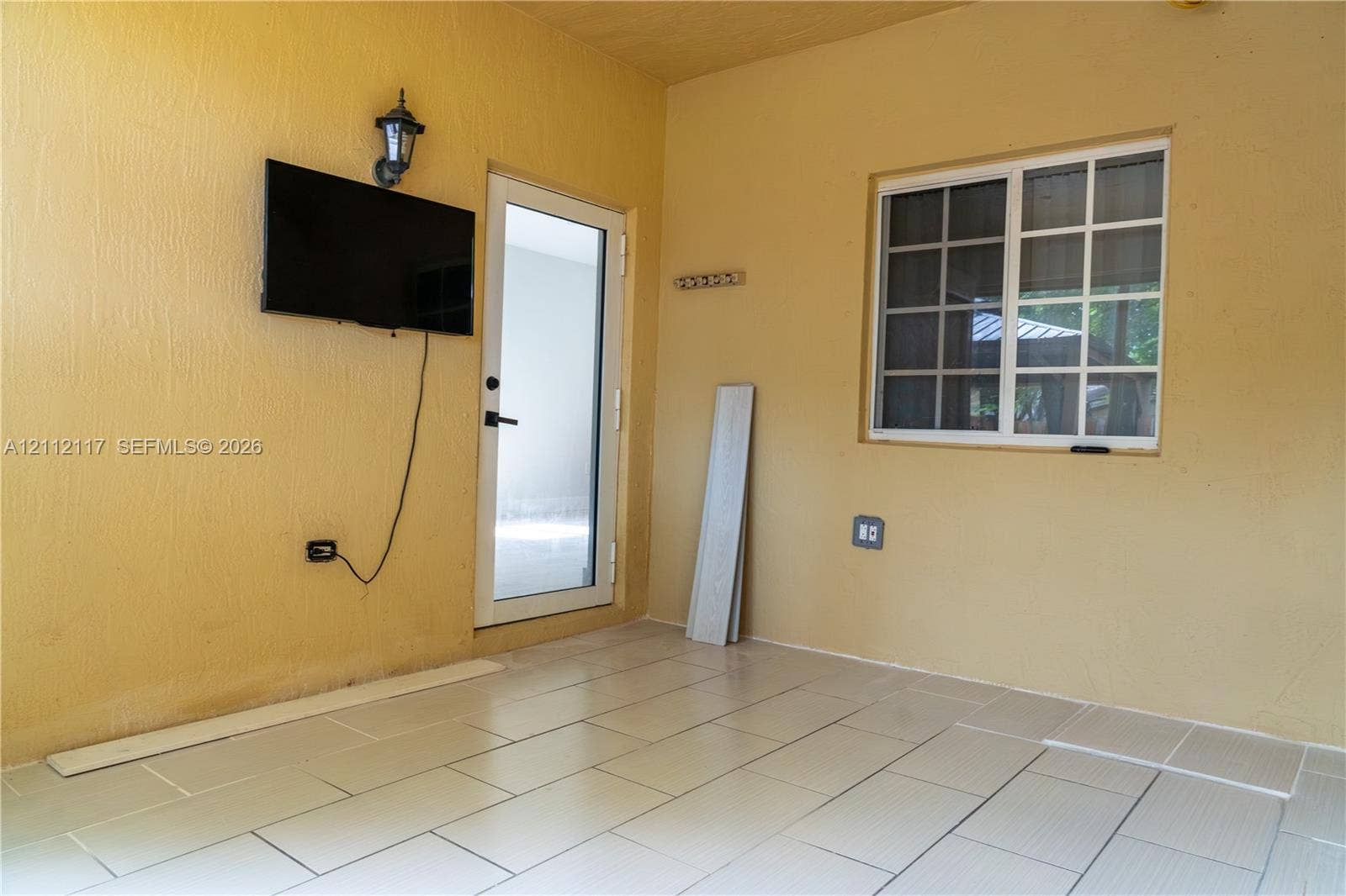 1209 NE 32nd Avenue, Homestead, Homestead, FL 33033 Covered outdoor area featuring neutral-toned tile flooring, a wall-mounted light fixture, and a glass-paneled entry door with a multi-pane window