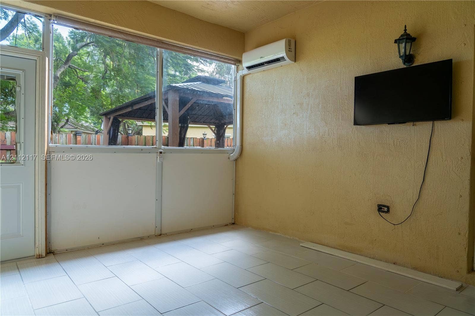 1209 NE 32nd Avenue, Homestead, Homestead, FL 33033 Enclosed patio featuring floor-to-ceiling screen panels, neutral ceramic tile flooring, a wall-mounted ductless air conditioning unit, and an exterior wall-mounted lantern light fixture