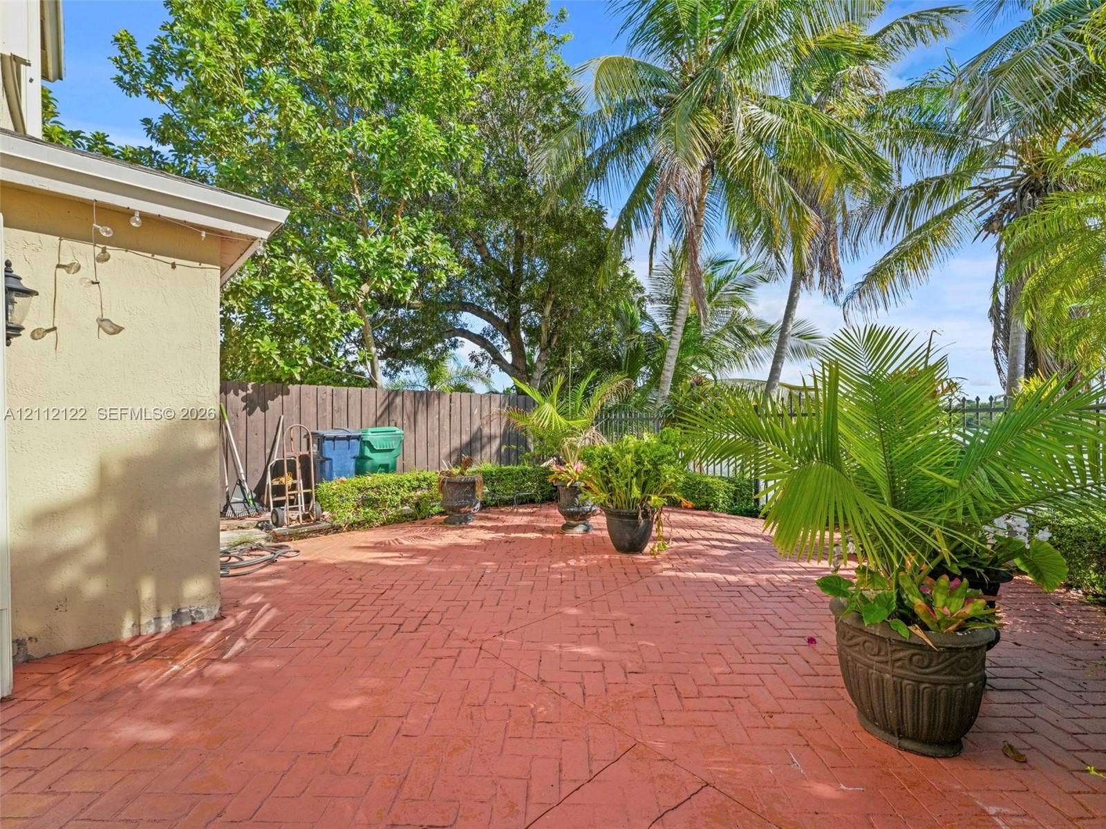 13990 SW 161st Terrace, Miami, FL 33177 Herringbone-patterned brick patio featuring a perimeter hedge, mature palm trees, and metal fencing