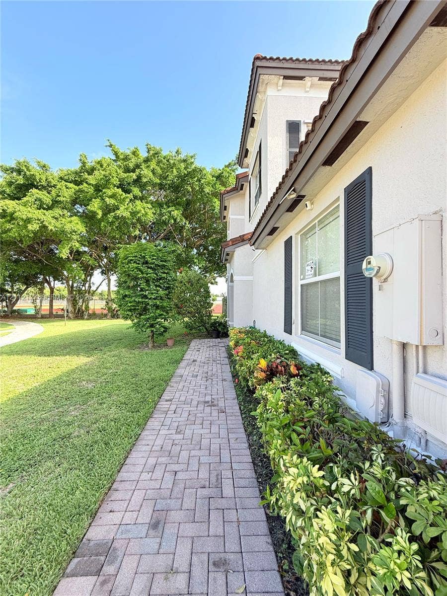 Stucco exterior featuring dark shutters, herringbone-pattern brick paver walkway, mature landscaping, and expansive green space