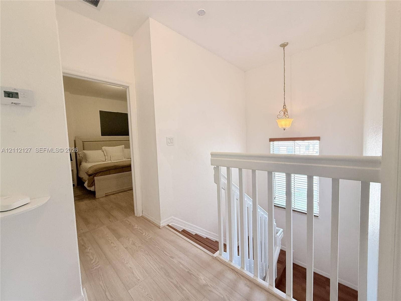 Hallway featuring wood-finish flooring, white spindle railing, a pendant light fixture, and horizontal window blinds