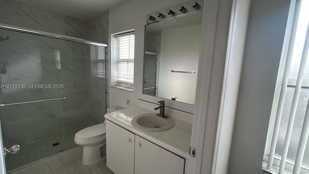 Bathroom featuring a stone-look countertop with an integrated basin, wall-mounted mirror, glass-enclosed shower with tiled surround, and metal faucet fixtures