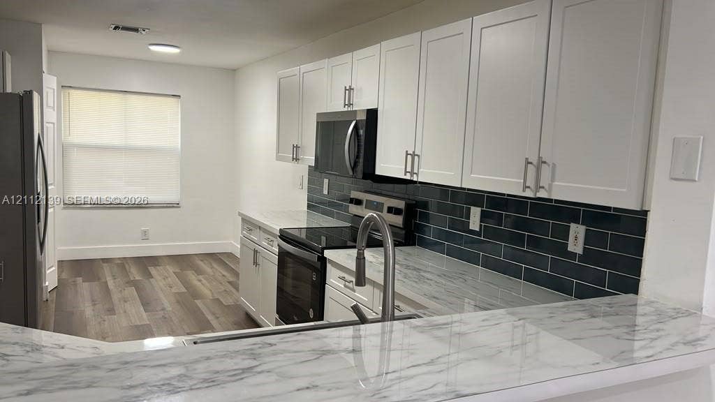Kitchen featuring white shaker-style cabinetry, a dark tile backsplash, stone-finish countertops, stainless steel appliances, and wood-finish flooring