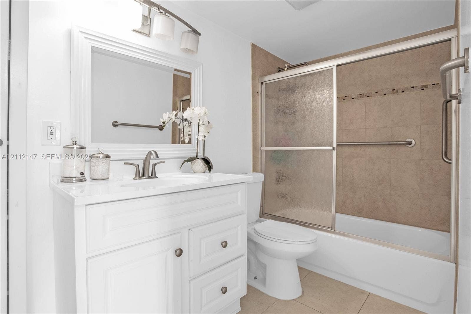 8331 Sands Point Boulevard, #C308, Tamarac, FL 33321 White cabinetry vanity with a countertop basin and multi-light fixture, framed mirror, and a tiled shower-tub combination with sliding glass doors