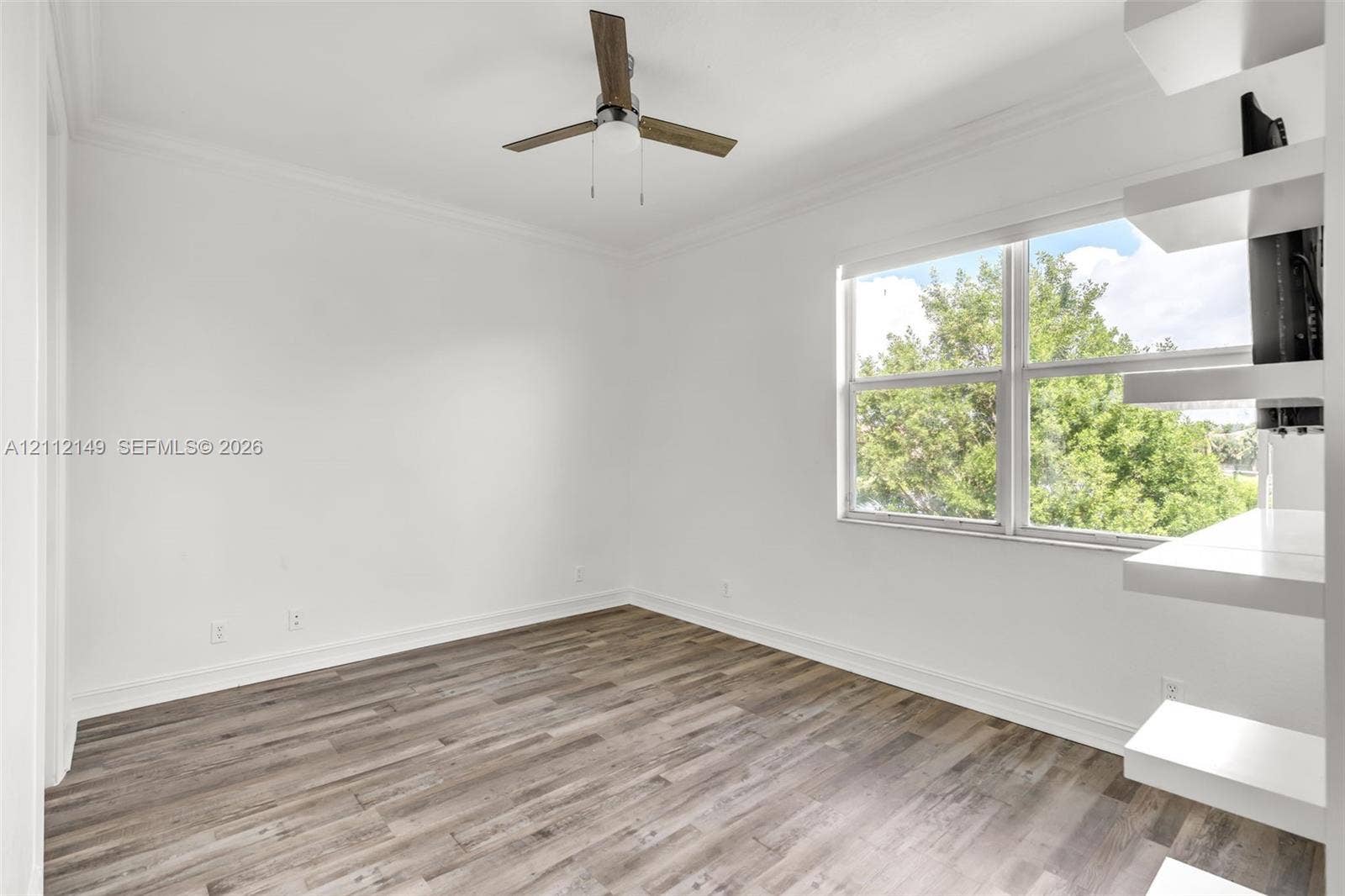 Wood-finish flooring, white built-in shelving, double-pane window, and modern ceiling fan with integrated lighting